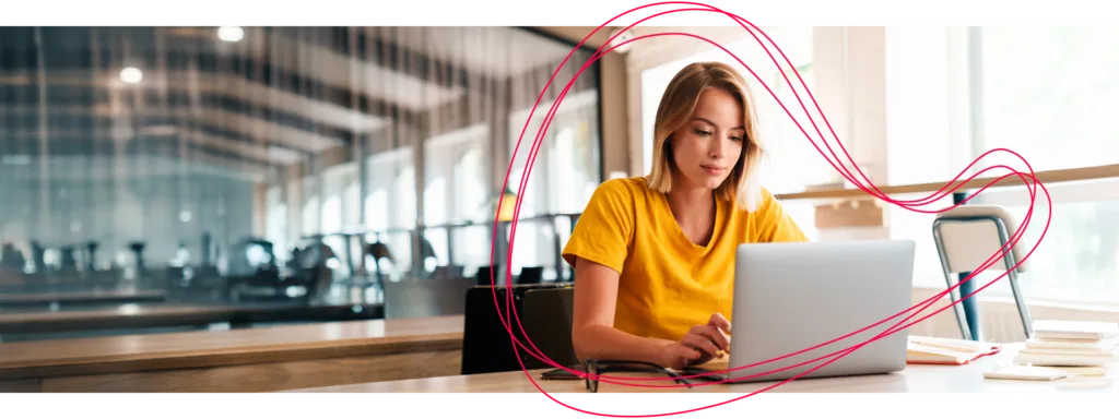 Eine Frau im gelben Shirt sitzt in einem B&uuml;ro an einem Laptop.