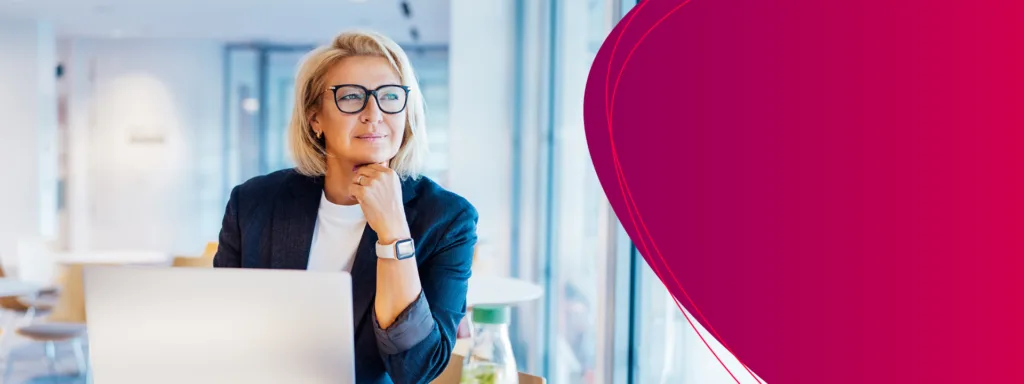 Frau mit Brille sitzt am Laptop und blickt nachdenklich aus dem Fenster.
