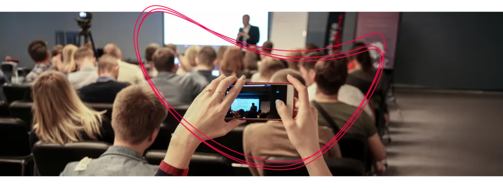 Publikum bei einem Vortrag; im Vordergrund filmt jemand die Bühne mit dem Smartphone.